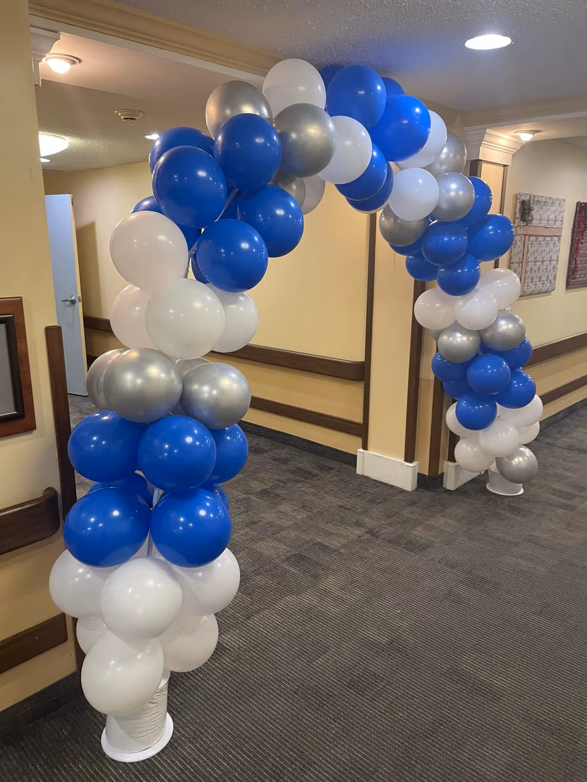 An archway made of blue, white, and silver balloons is set up in a hallway with beige walls and carpeted floor inside a senior living facility.