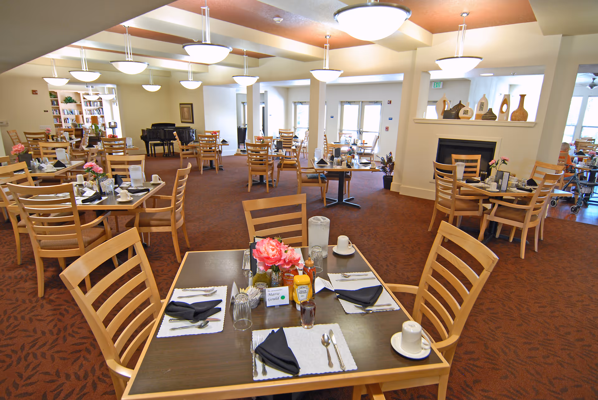 A spacious dining room in an assisted living facility with multiple wooden tables and chairs arranged neatly. Each table is set with placemats, napkins, utensils, cups, and condiments. The room has warm lighting with ceiling fixtures, a carpeted floor, and decorative vases on a shelf above a fireplace. A grand piano and bookshelves are visible in the background.