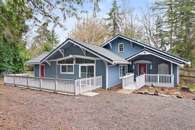 Exterior view of a single-story blue house with white trim and a gray roof, surrounded by trees. The house features a white railing along a ramp and porch area, with two red doors visible. The ground in front is covered with gravel and some patches of grass.