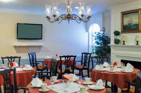 A senior living dining room with round tables, red tablecloths, chairs, chandelier, and fireplace.