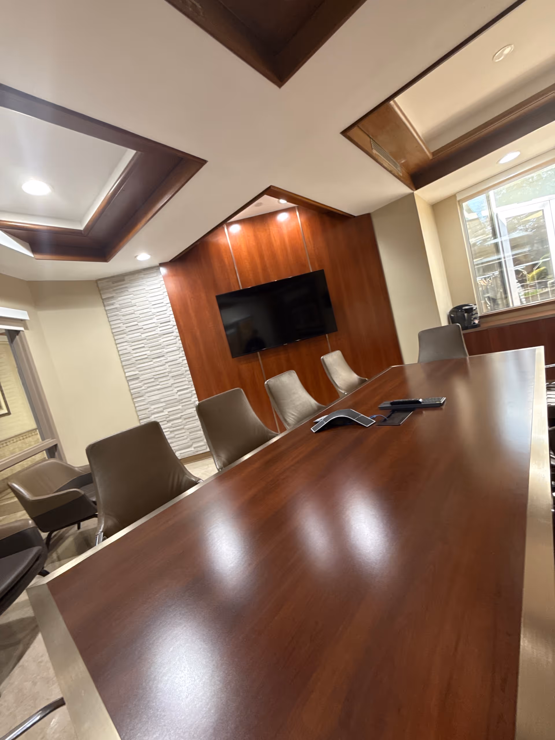 A modern conference room with a long wooden table surrounded by several brown leather chairs. The room features a wall-mounted flat screen TV on a wooden panel wall, recessed ceiling lights, and a window letting in natural light. A conference phone and remote control are placed on the table.