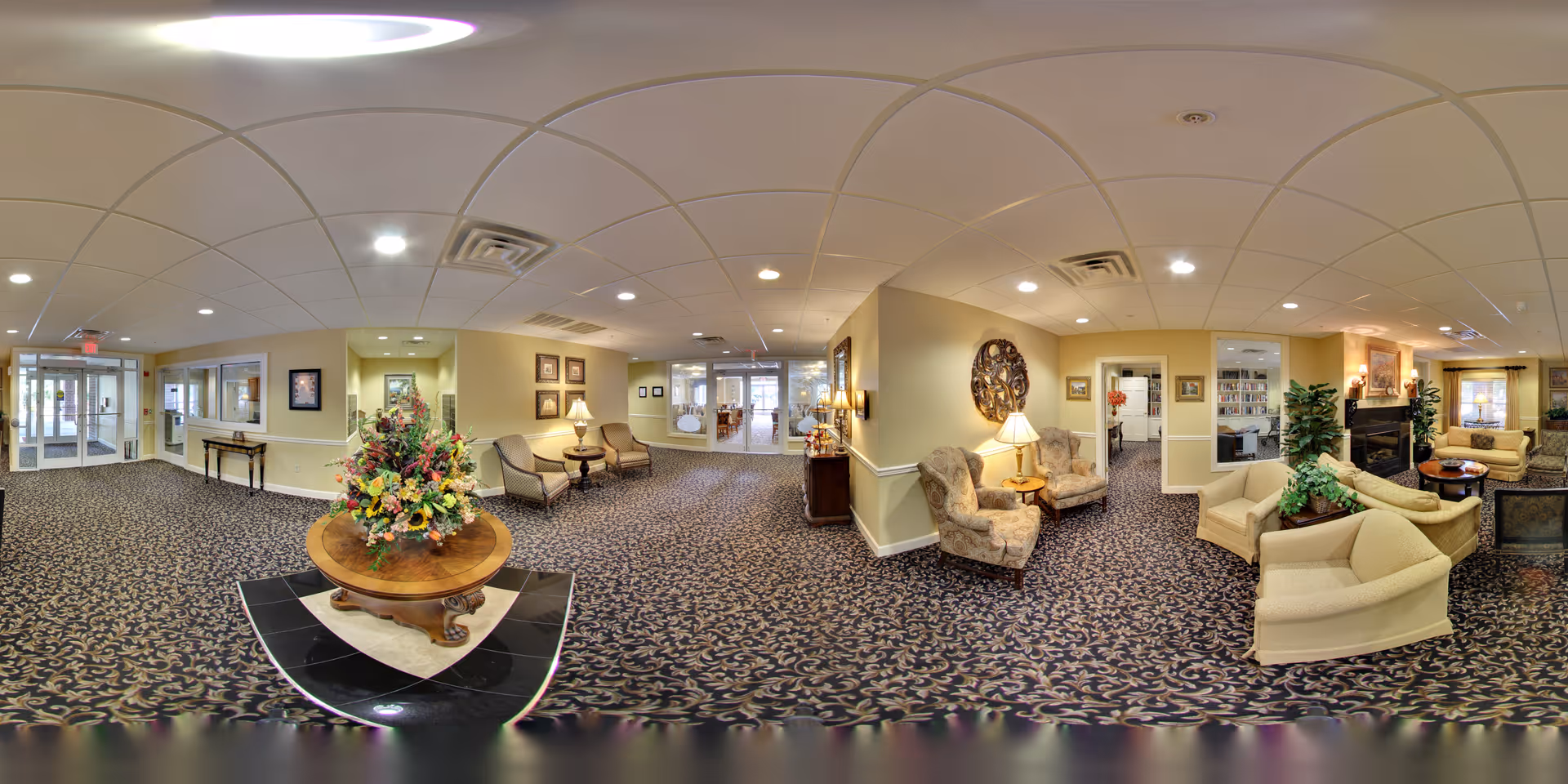 A spacious and well-lit senior living facility common area with patterned carpet, beige walls, and ceiling lights. The room features multiple seating arrangements including armchairs and sofas, a round wooden table with a large floral arrangement in the center, lamps, framed artwork on the walls, and a fireplace with plants and decorative items. There are glass doors and windows leading to other rooms and the entrance area.