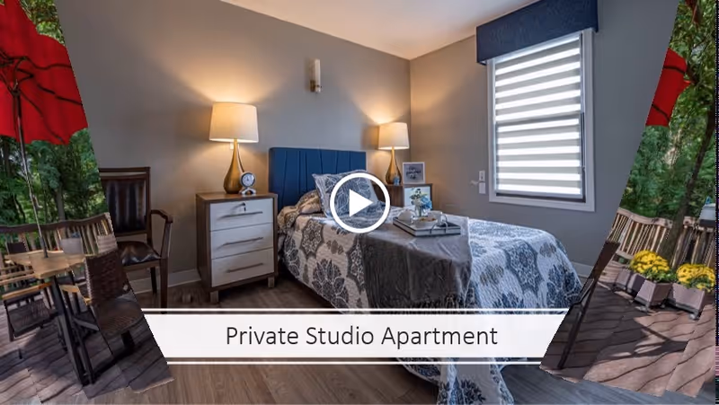 A cozy private studio apartment bedroom with a single bed covered in a patterned bedspread and a gray throw blanket. There are two bedside tables with lamps on each side of the bed, a chair, and a window with a modern blind. The room has wooden flooring and neutral-colored walls.