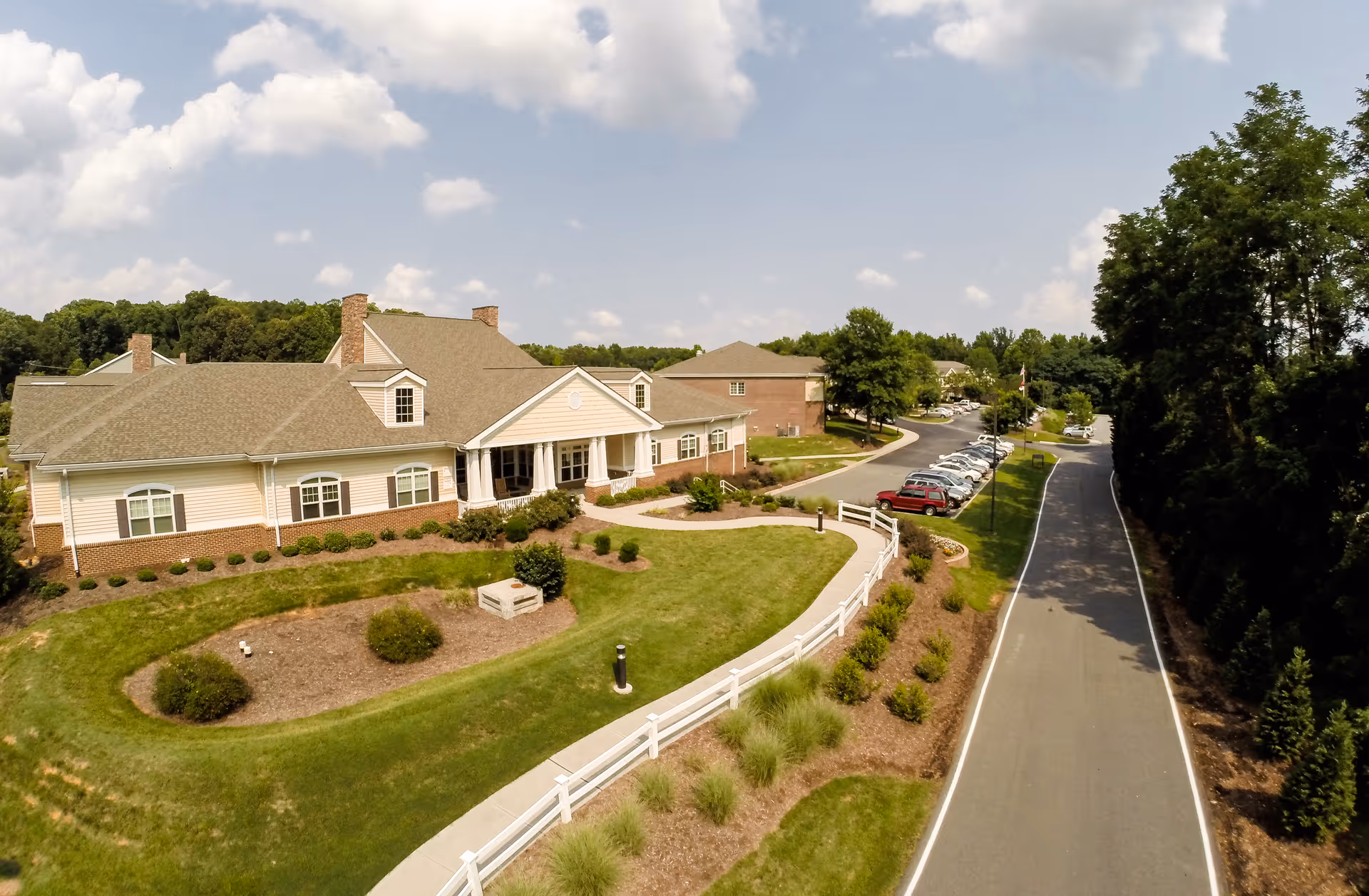 Aerial view of Piedmont Crossing Retirement Community in Thomasville, NC, showing a large building with beige siding and brick accents, surrounded by well-maintained lawns, shrubs, and a white fence. A paved road runs alongside the property with parked cars and trees lining the road.