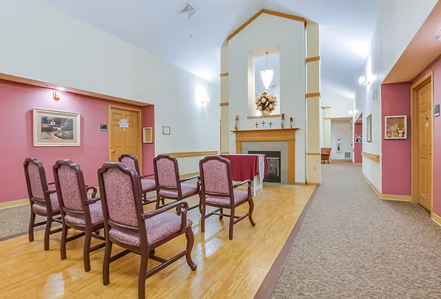 Interior view of a senior living facility chapel area with six upholstered chairs arranged in two rows facing a small altar with a floral arrangement and three crosses above a fireplace. The walls are painted white and pink, with wooden trim, and a hallway extends to the right with additional seating and artwork on the walls.