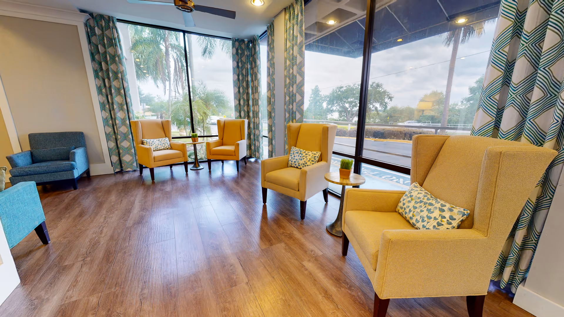 A bright and spacious seating area with large floor-to-ceiling windows showing outdoor greenery. The room has wooden flooring, patterned curtains, and several armchairs in yellow and blue with small side tables holding decorative plants.