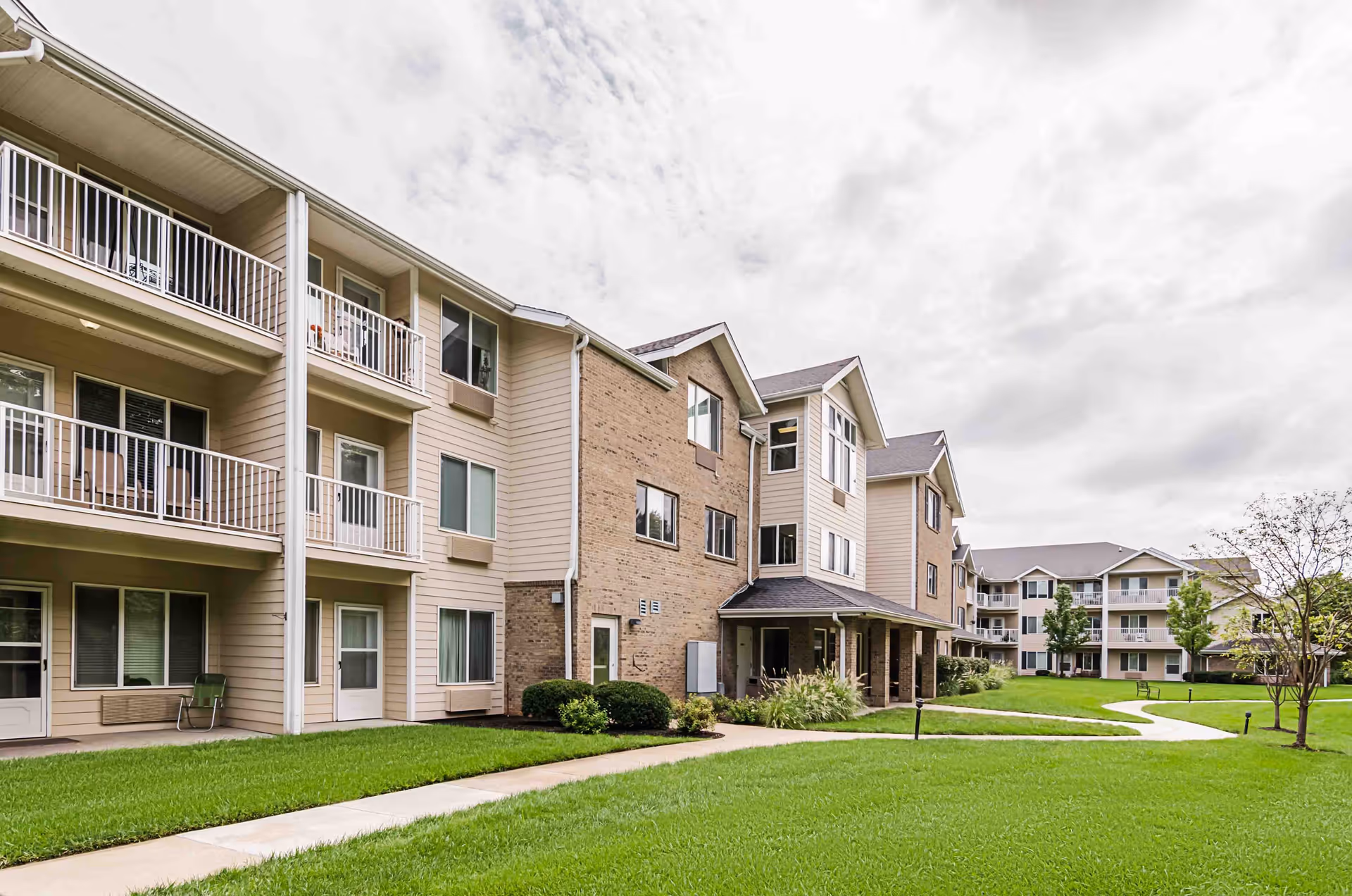 Exterior view of Holiday Greenwood Terrace senior living facility showing multi-story buildings with balconies, well-maintained green lawns, paved walkways, and some trees under a cloudy sky.