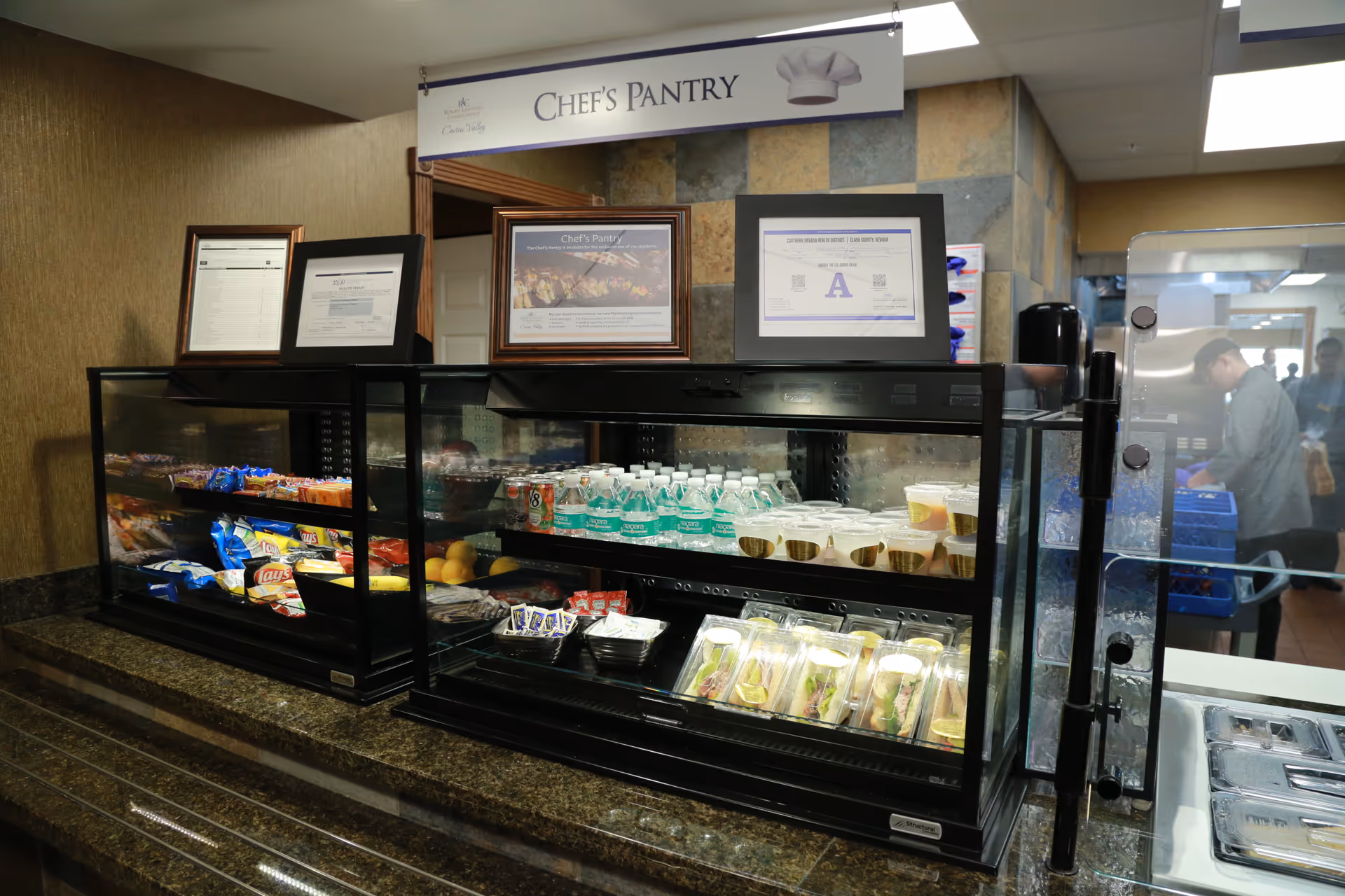 A food display counter labeled 'Chef's Pantry' featuring a variety of snacks including chips, bottled water, pre-packaged sandwiches, and cups of pudding or gelatin. Behind the counter, framed certificates and menus are displayed on the wall. A person is visible in the background working in the kitchen area.
