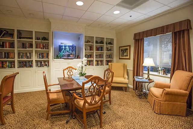 A cozy common area with a wooden table and six chairs in the center, surrounded by built-in bookshelves filled with books. There is a large aquarium set into the wall above the bookshelves. Two upholstered armchairs and a small side table with a lamp are positioned near a window with brown curtains. The room has a patterned carpet and a ceiling with recessed lighting.