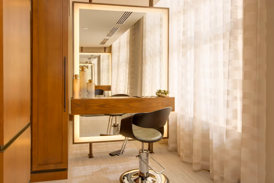 A salon-style vanity station with a backlit mirror, wooden counter and a black swivel chair beside sheer curtains.
