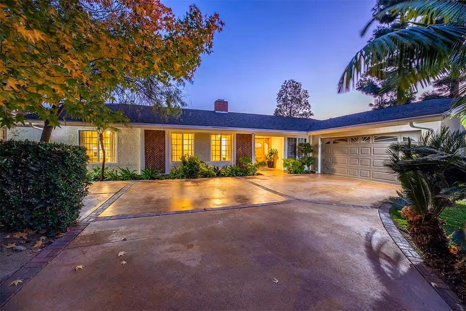 Exterior view of a single-story house at dusk with warm lights glowing from the windows. The driveway is spacious and clean, surrounded by well-maintained greenery including bushes, trees with autumn leaves, and palm plants. The house features a garage on the right side and a front entrance with plants near the door.