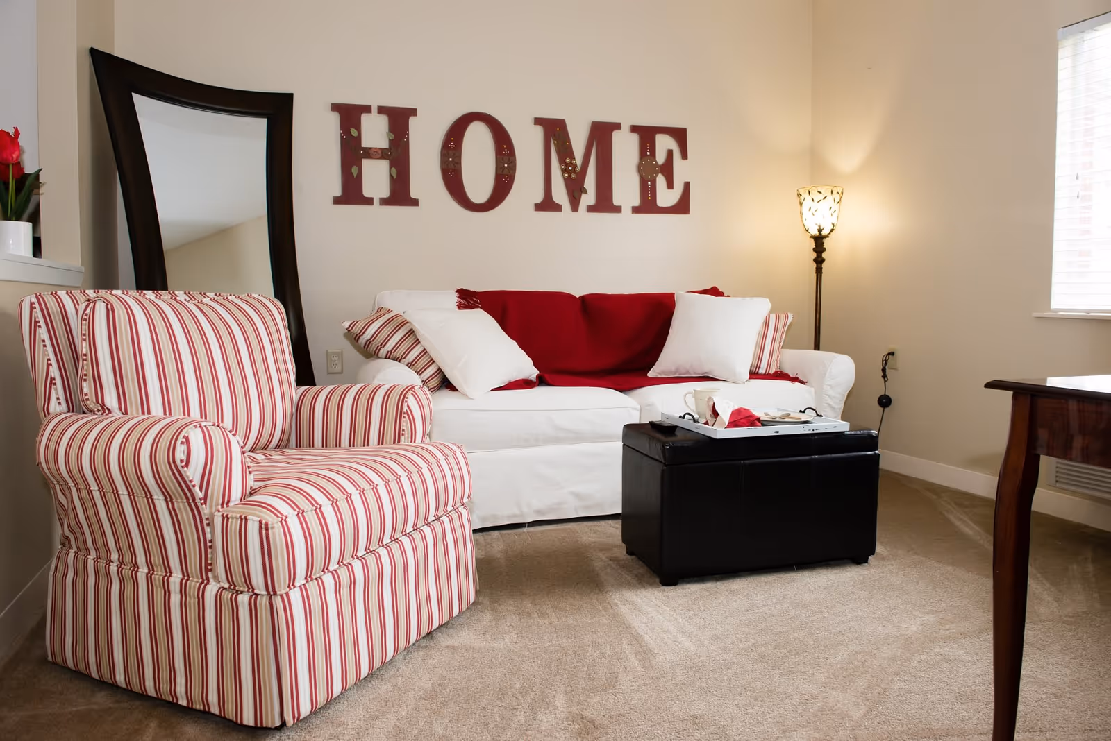 A cozy living room with a white sofa adorned with red and white pillows and a red throw blanket. A striped armchair with red and white stripes sits to the left. Behind the sofa, large letters spelling 'HOME' are mounted on the wall. A tall floor lamp with a decorative shade is in the corner, and a large mirror leans against the wall. A black ottoman with a tray on top is in front of the sofa, and a wooden table is partially visible on the right. The room has beige carpet and light-colored walls with a window letting in natural light.