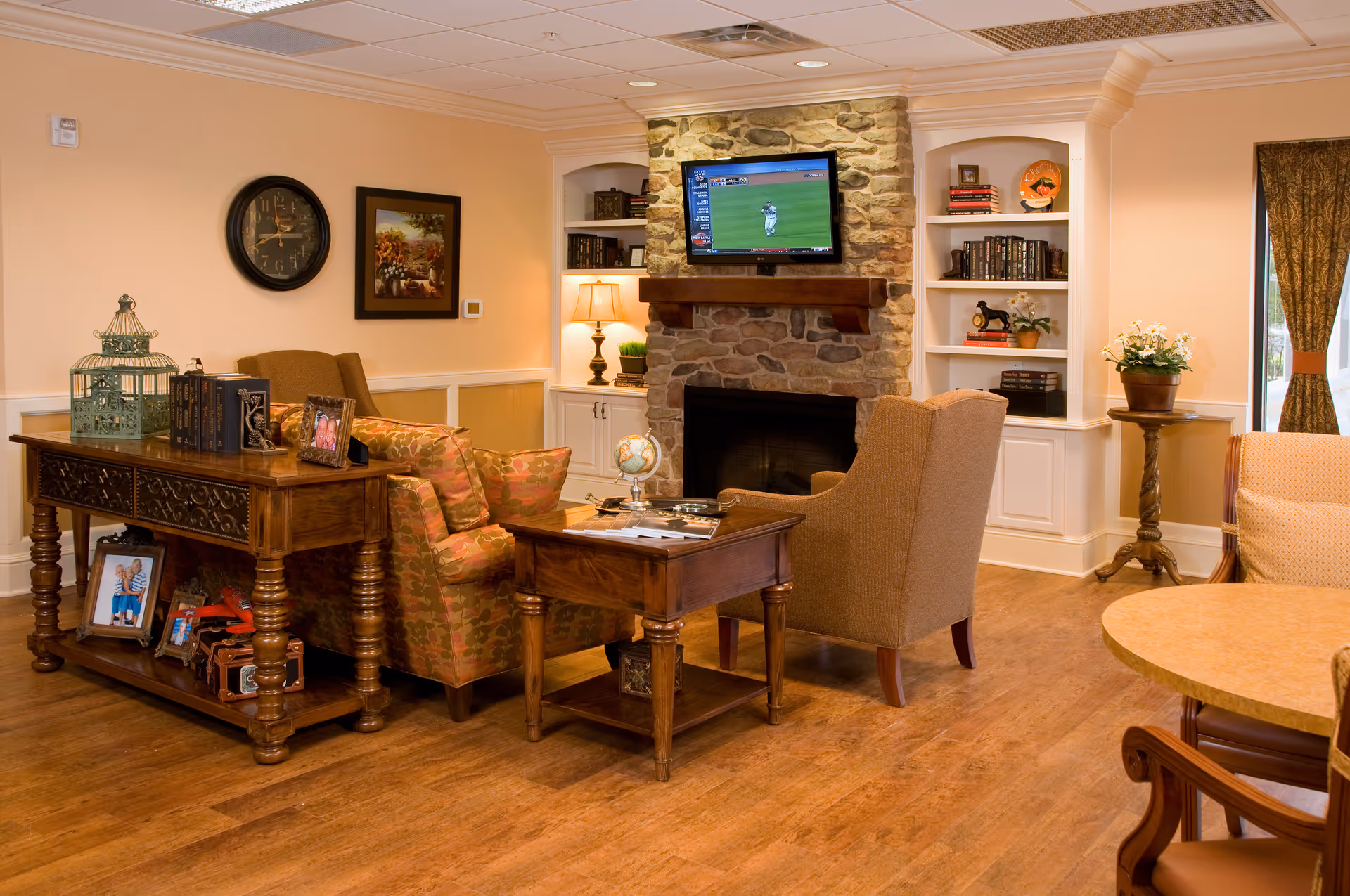 Cozy senior living lounge with armchairs, wooden tables, a stone fireplace with a wall-mounted TV, and built-in bookshelves.