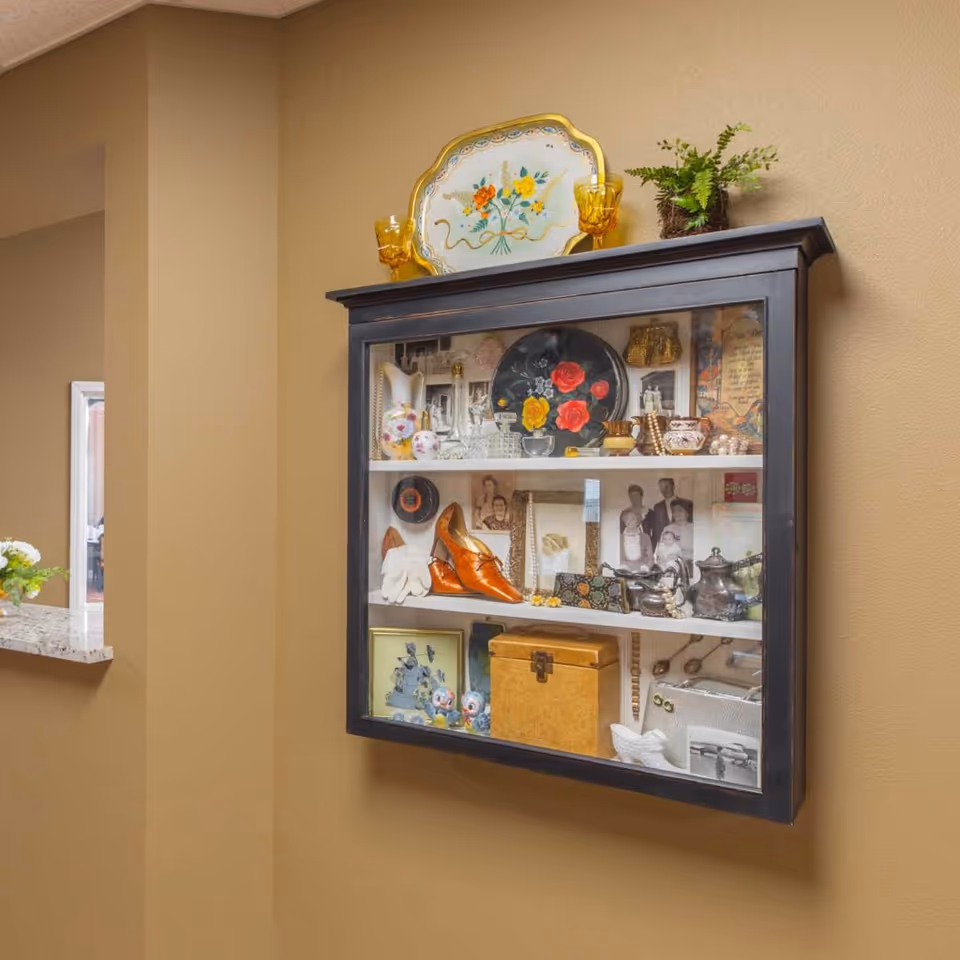 Glass-front wall display cabinet filled with vintage decorative items, photos and a plant mounted on a beige interior wall.