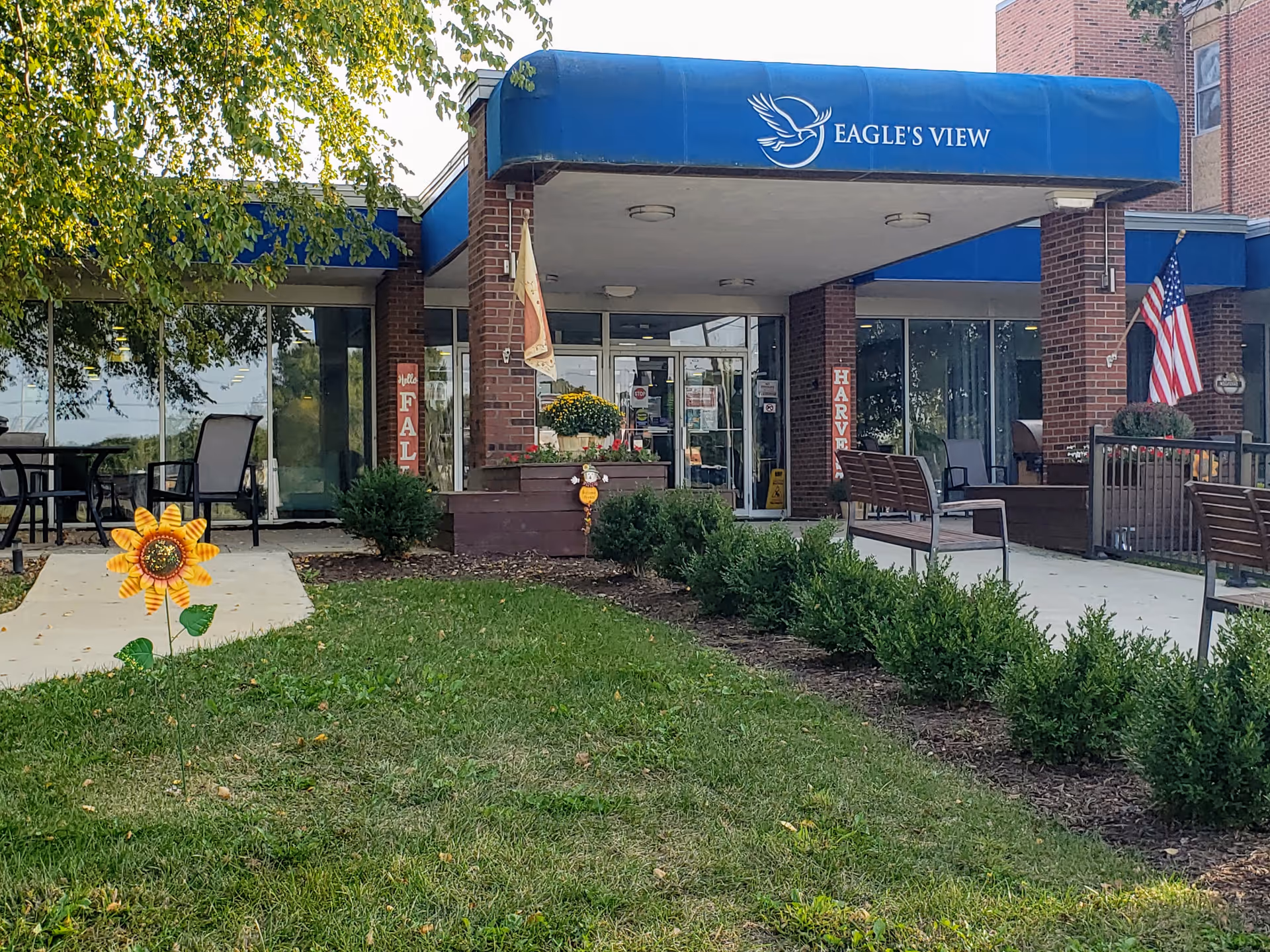 Entrance to Eagle's View supportive living facility with a blue awning bearing the name, glass doors, benches, planters, and a grassy front lawn.