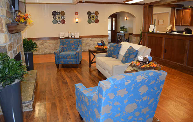 A cozy nursing home lobby area with wooden flooring, a stone fireplace on the left, and comfortable seating including a beige sofa with blue floral cushions and two blue floral armchairs. There are decorative plants, a wooden reception desk on the right, and wall decorations with a warm, inviting ambiance.