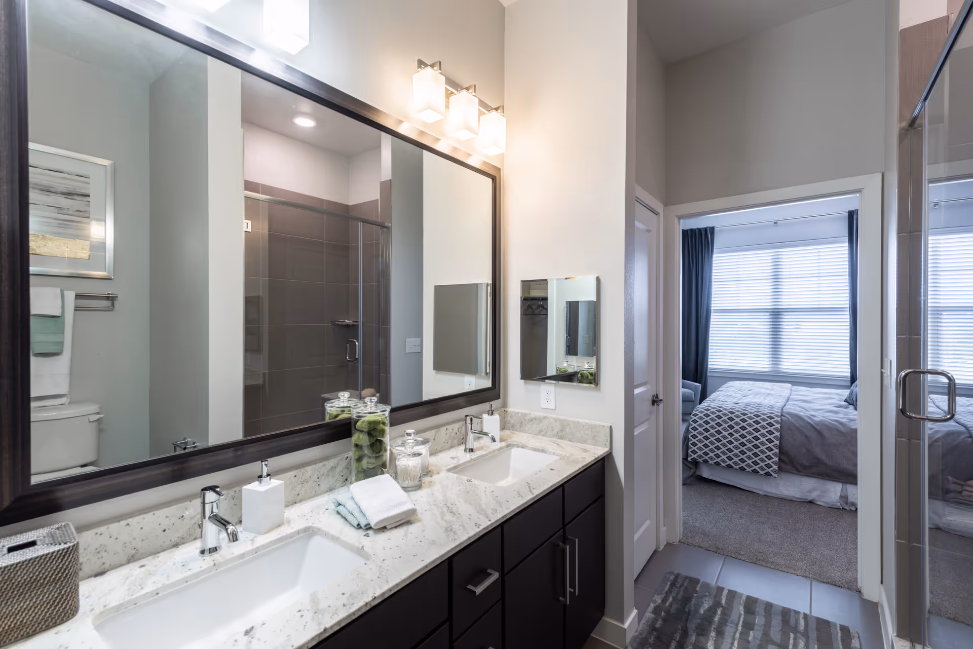 Modern bathroom with a double sink vanity featuring a marble countertop, two rectangular sinks, and a large mirror above. The bathroom has a glass-enclosed shower with gray tiles and a toilet with towels hanging on a rack above it. Through an open doorway, a bedroom with a bed, patterned blanket, and large window with blinds is visible.