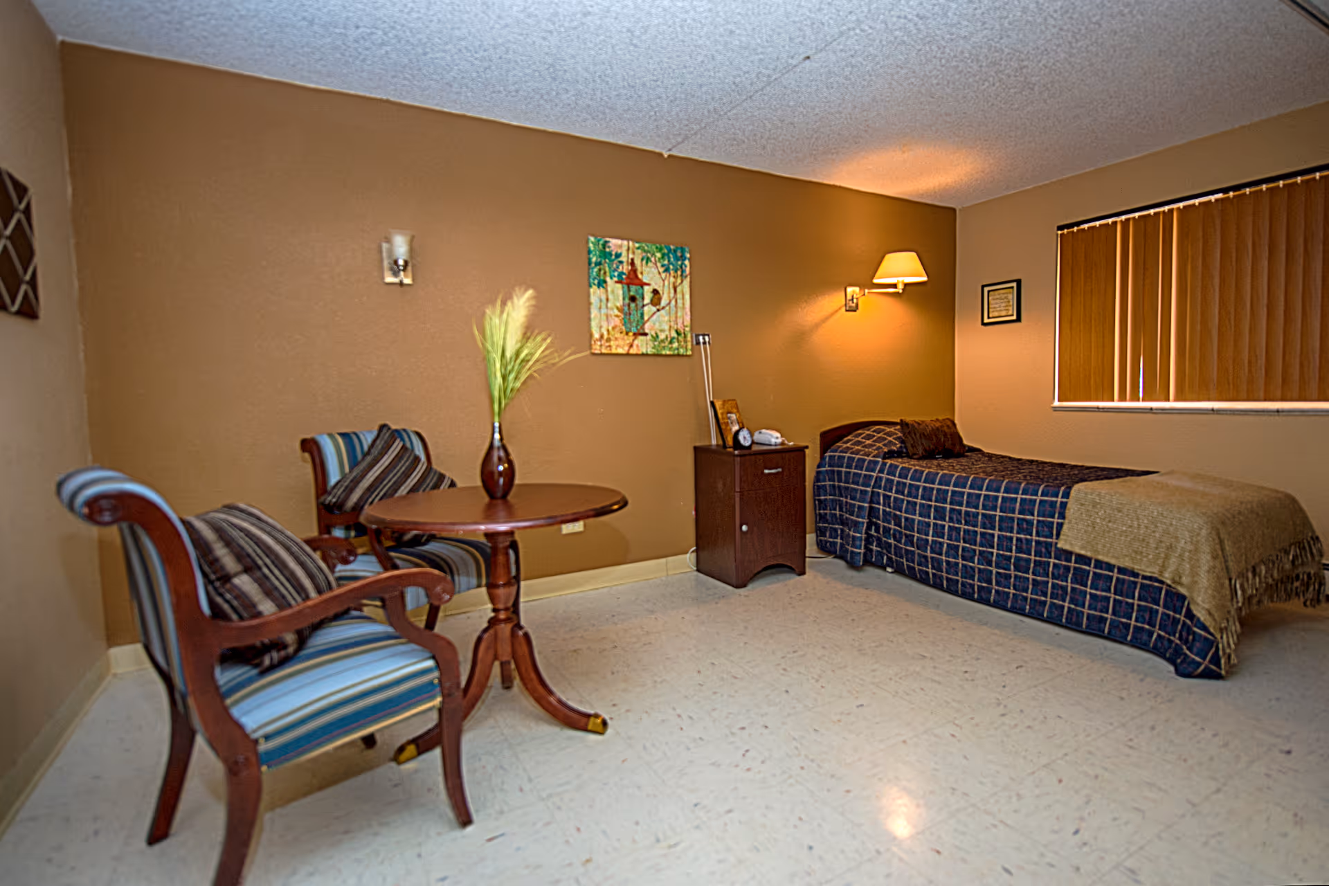 A simple bedroom with a single bed covered in a dark blue checkered blanket and a brown throw. Next to the bed is a small wooden nightstand with a lamp, clock, and phone. On the left side of the room, there are two wooden chairs with striped cushions and a round wooden table with a vase holding decorative grass. The walls are painted brown, and there is a window with closed vertical blinds. A colorful painting hangs on the wall above the nightstand.