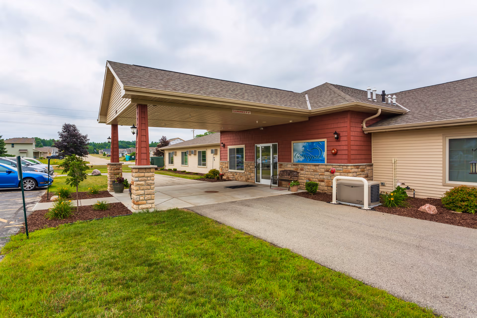 Front entrance of a single-story senior living facility with a covered drive-up canopy, stone pillars, and landscaped lawn.