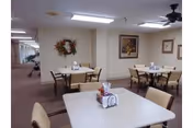 Interior view of a senior living facility dining area with several square tables and chairs arranged neatly. Each table has a centerpiece with napkins and condiments. The walls are decorated with framed artwork and a floral wreath. The room is well-lit with ceiling lights and has a carpeted floor.