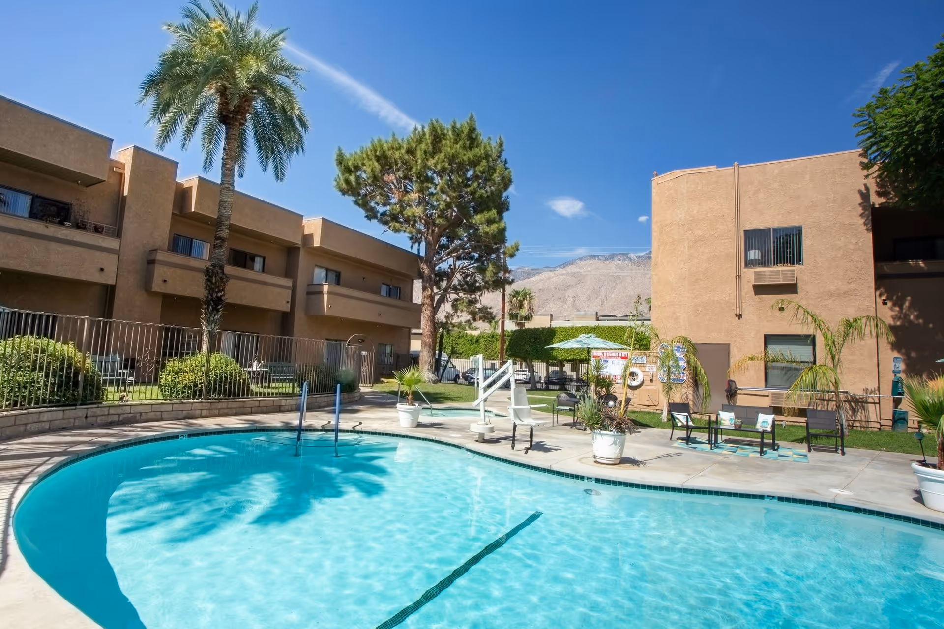 Outdoor courtyard featuring a swimming pool, lounge chairs, potted plants, and apartment-style assisted living buildings under a clear blue sky.