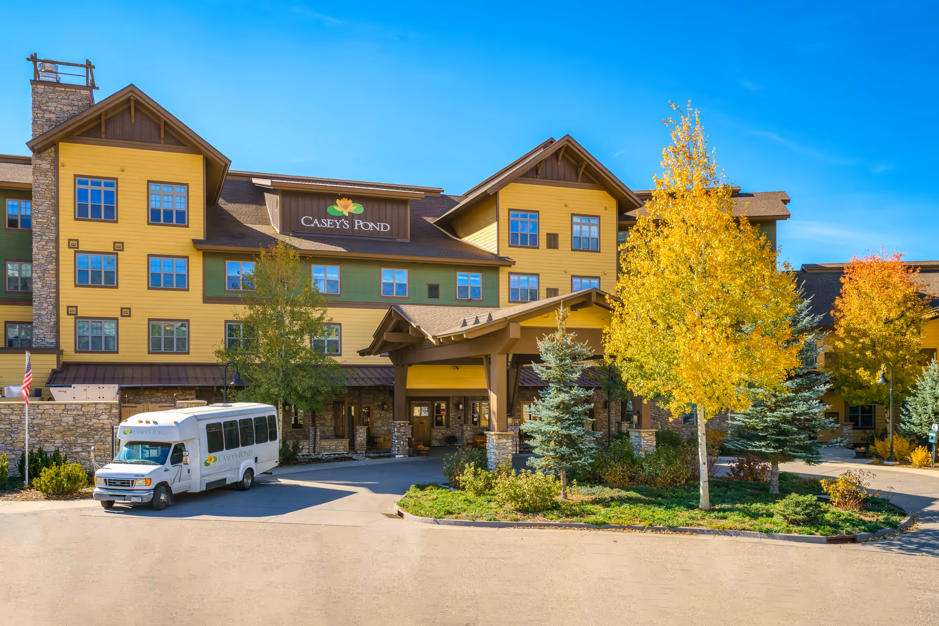 Front exterior of a yellow multi-story senior living building labeled "Casey's Pond" with a shuttle van parked at the entrance and autumn trees in front.