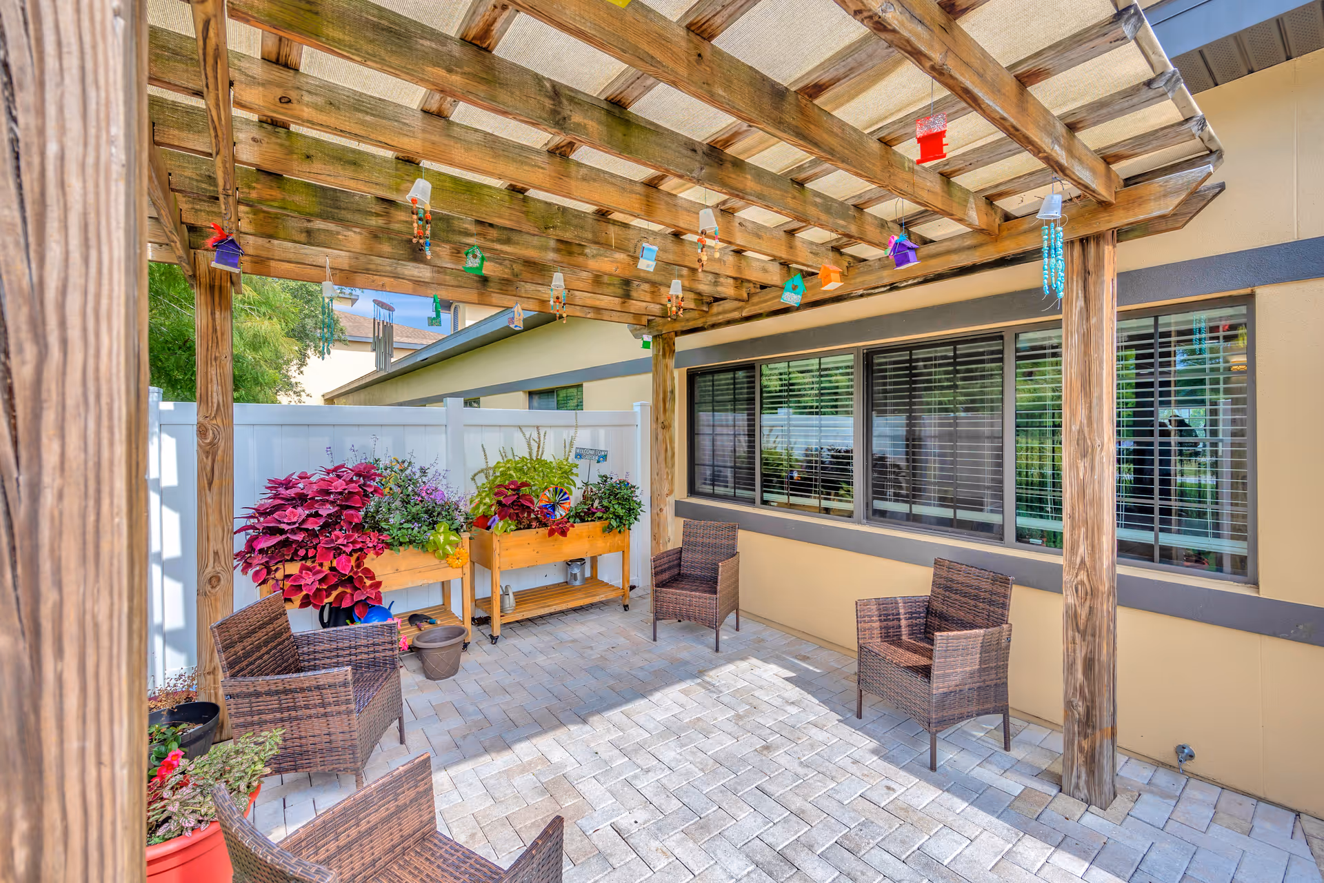 Outdoor patio area with a wooden pergola overhead, decorated with hanging colorful birdhouses and wind chimes. The patio has a paved floor and four wicker chairs arranged around the space. There are two wooden planter boxes filled with vibrant flowers and plants against a white fence. The building wall with large windows and closed blinds is visible on one side.