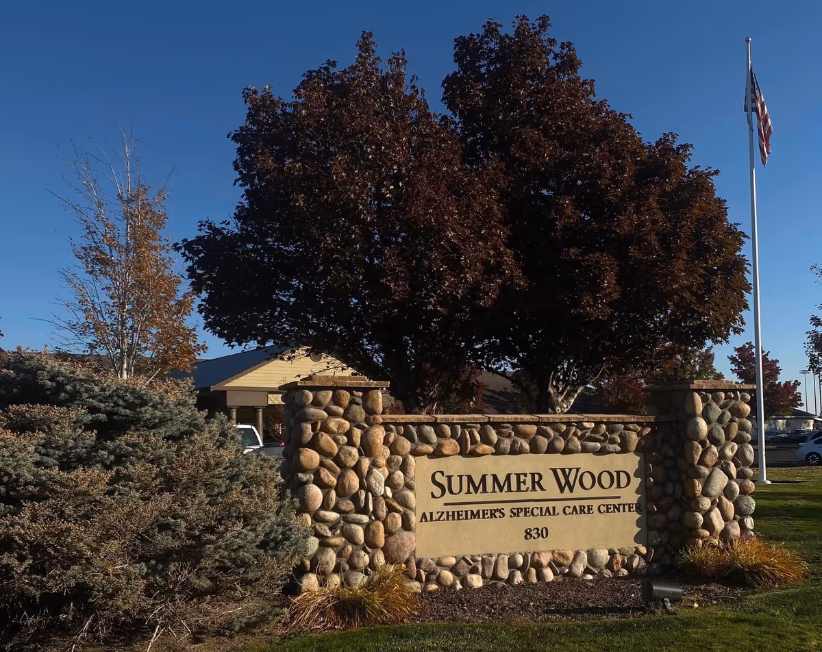 Stone sign for Summer Wood Alzheimer's Special Care Center with the number 830, surrounded by bushes and trees under a clear blue sky with an American flag on a flagpole in the background.
