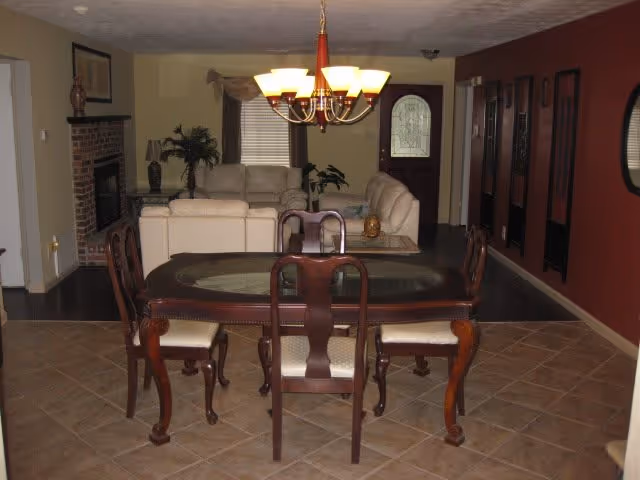 Open-plan dining area with a wooden table and six chairs, a chandelier overhead, and a living room with sofas, fireplace, and front door in the background.