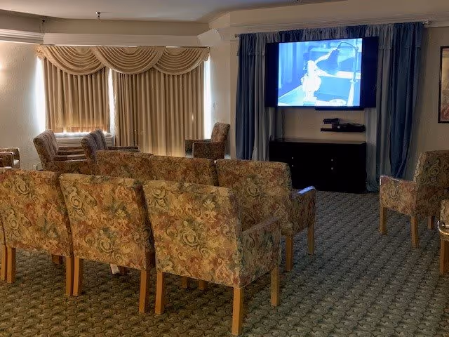 A small theater or media room with several rows of floral-patterned upholstered chairs facing a wall-mounted flat screen TV. The room has carpeted flooring, beige walls, and windows with beige curtains and valances. Blue curtains frame the TV area.