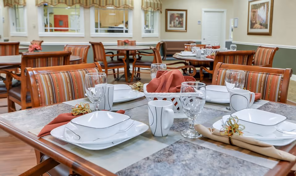 A neatly set dining table with white dinnerware, glassware, cloth napkins and striped chairs in a senior living dining room.