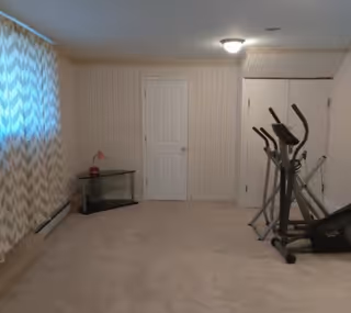 A carpeted room with beige walls and a ceiling light. There is a white door in the center of the back wall, a small glass table with a red lamp on the left side near a window with patterned curtains, and an elliptical exercise machine on the right side near a set of double doors.