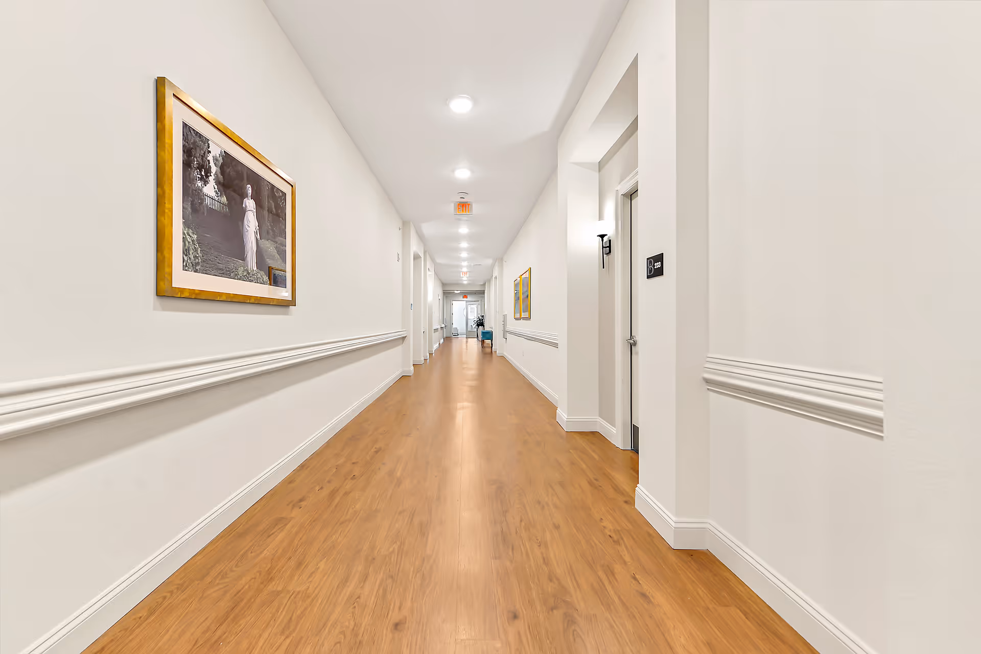 Long, well-lit hallway with wooden flooring and white walls featuring framed artwork. Several doors line the hallway, and ceiling lights illuminate the space evenly.