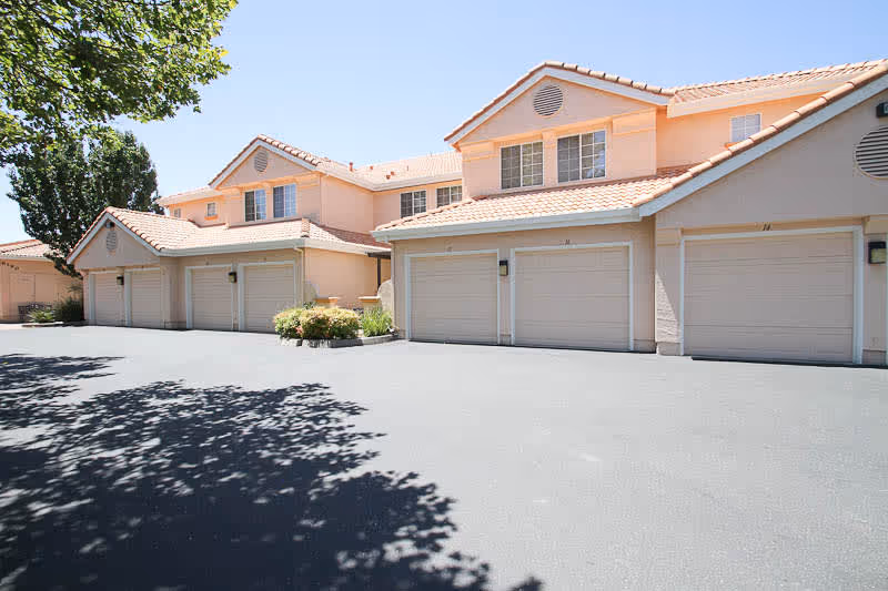 Exterior view of a senior living facility building with multiple attached garages, peach-colored walls, and tiled roofs under a clear blue sky. There are trees casting shadows on the paved driveway in front of the garages.