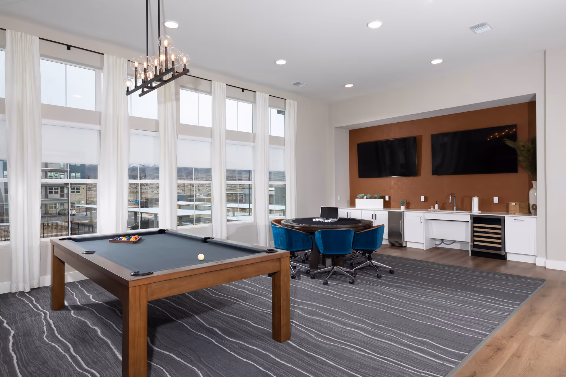 Bright communal game room with a pool table, round poker table with blue chairs, two wall-mounted TVs, and a kitchenette along the back wall.