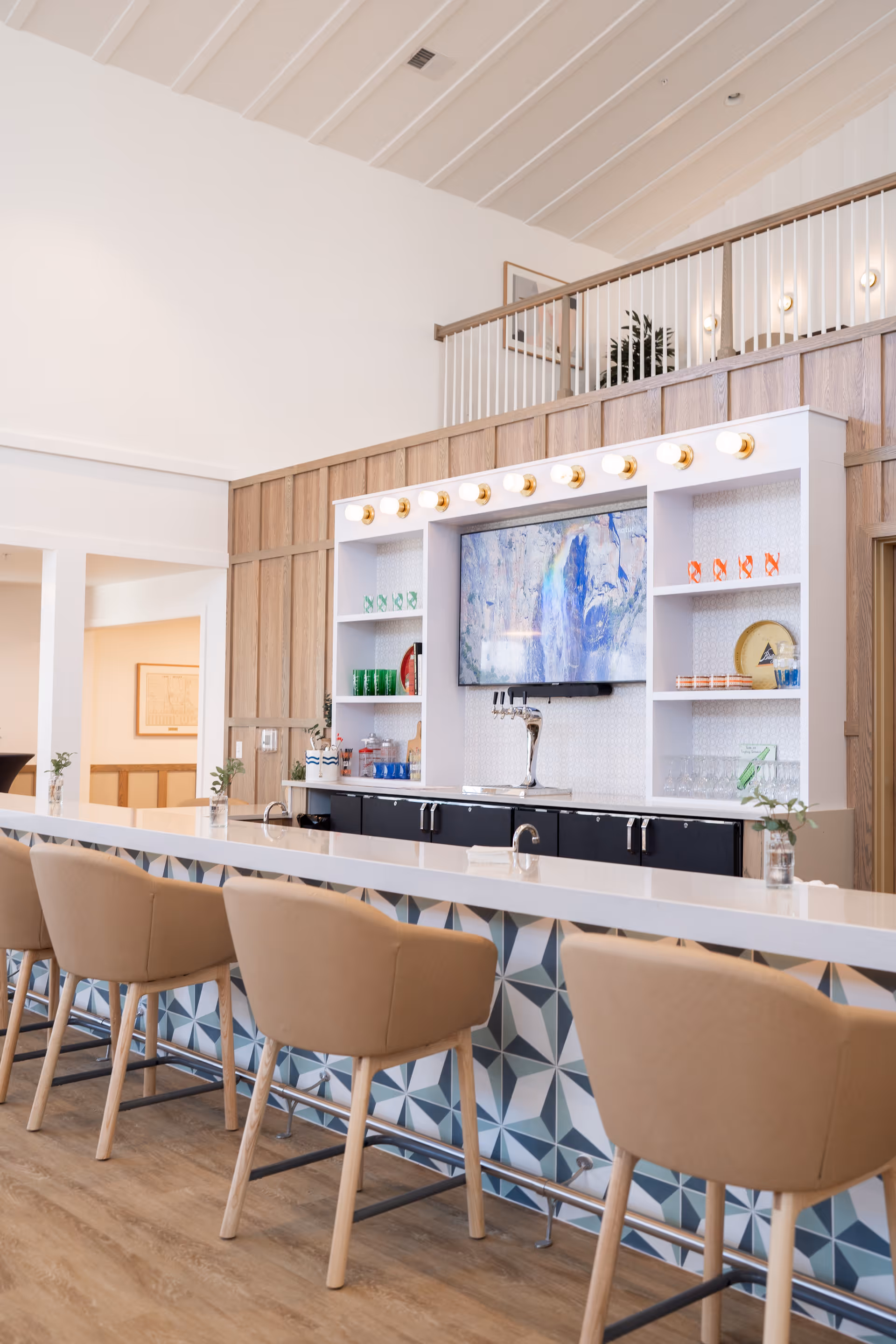 Bright communal bar area with a long white counter, beige barstools, a TV mounted in shelving, and decorative items behind the counter.