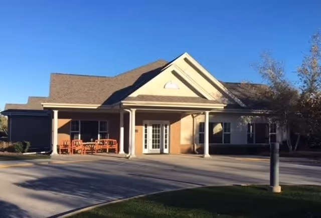 Front exterior view of a single-story assisted living facility building with a covered entrance supported by columns, outdoor benches, and a driveway in front. Trees and grass are visible around the building under a clear sky.