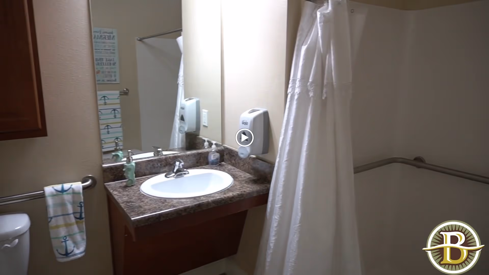 A bathroom with a sink and countertop, a mirror above the sink, a soap dispenser mounted on the wall, a towel hanging on a grab bar next to a toilet, and a shower area with a white curtain and grab bars.