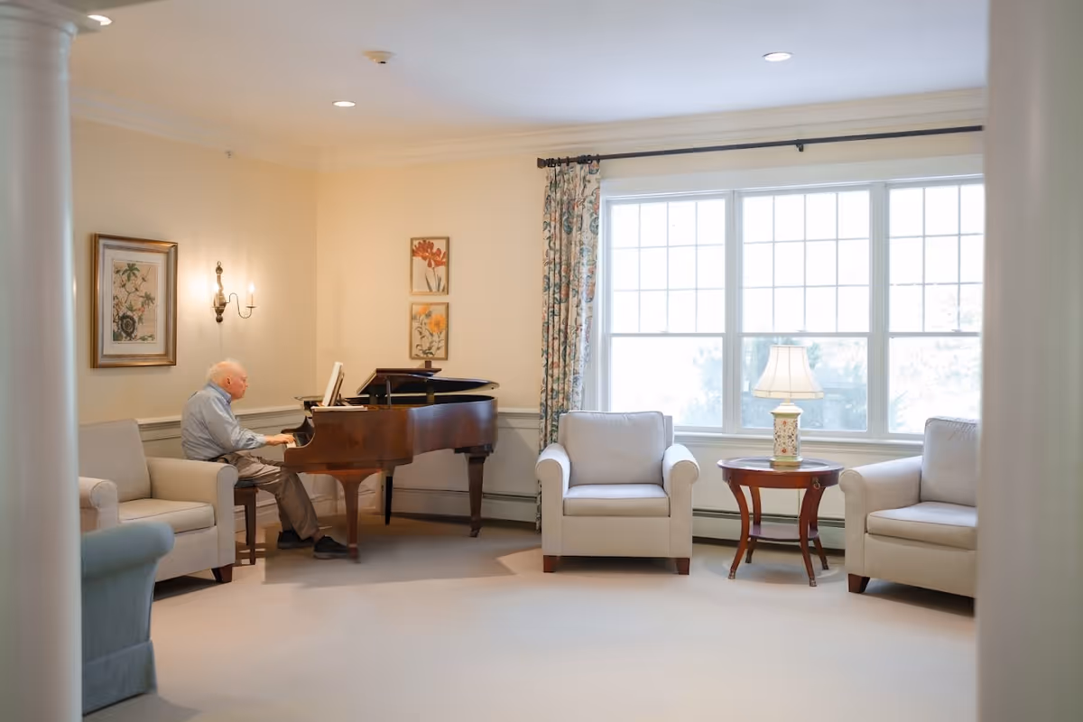 An elderly man playing a grand piano in a bright, cozy living room with beige walls and carpet. The room features three beige armchairs, a wooden side table with a decorative lamp, floral curtains, and framed botanical artwork on the walls.