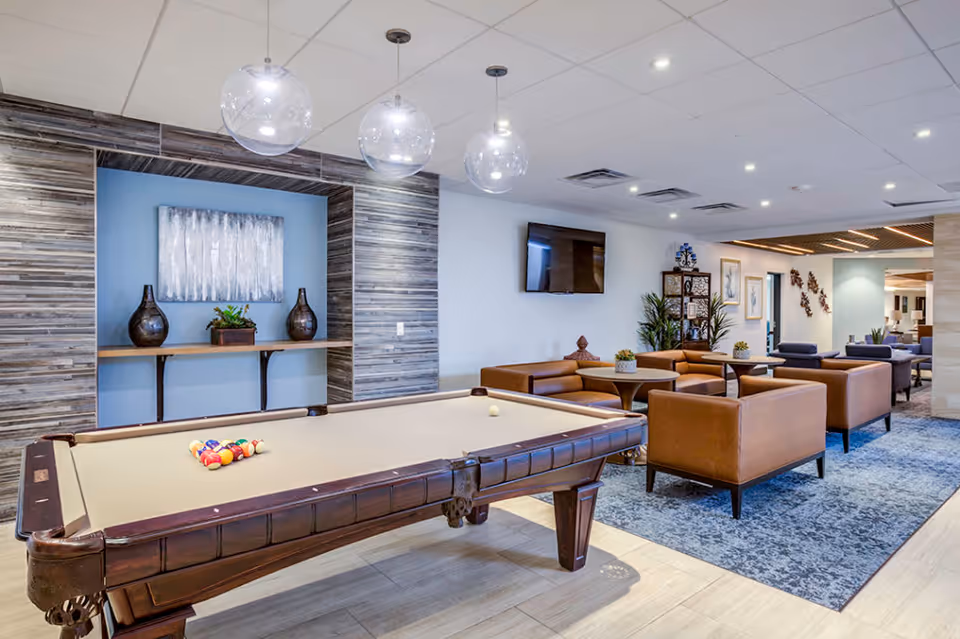 A modern lounge area featuring a pool table with billiard balls set up, surrounded by brown leather armchairs and small round tables. The room has a blue and gray color scheme with a decorative wall niche holding vases and a plant, a wall-mounted TV, and contemporary lighting fixtures hanging from the ceiling.