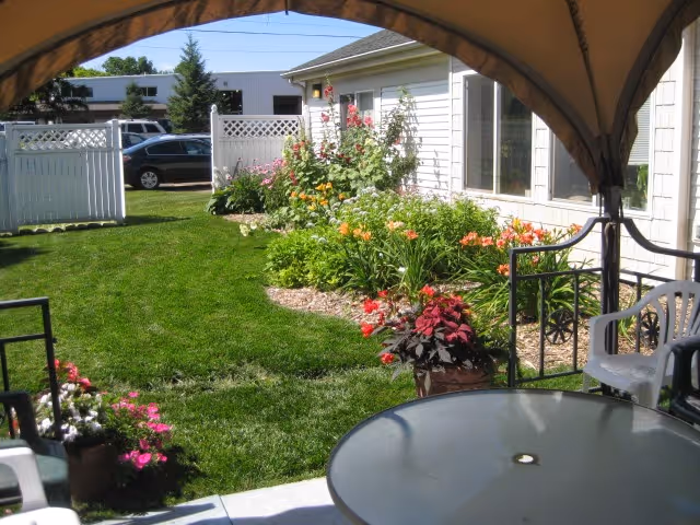 View of a well-maintained garden area with green grass, colorful flowers, and shrubs along the side of a light-colored building. The image is taken from under a canopy with a round glass table and white plastic chairs in the foreground.