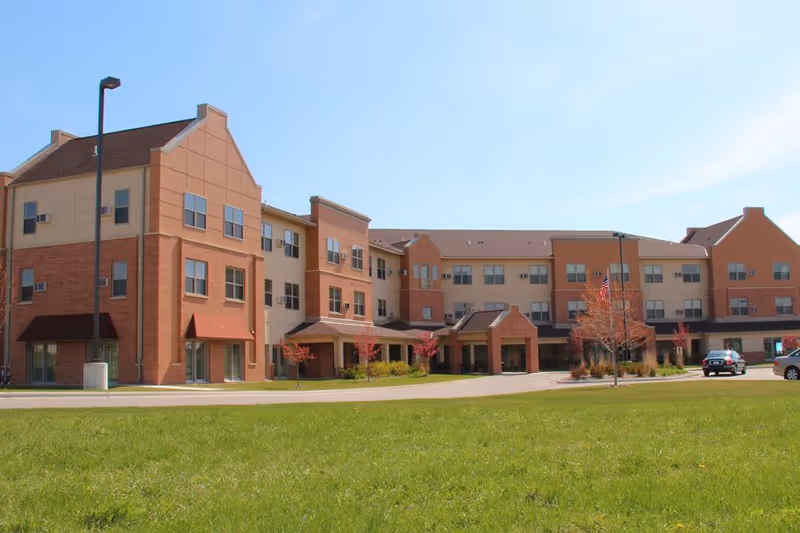 Exterior view of a three-story senior living facility building with a mix of brick and beige siding, multiple windows, and a covered entrance. There is a green lawn in the foreground and a few small trees with red leaves near the building. A car is parked near the entrance under a clear blue sky.