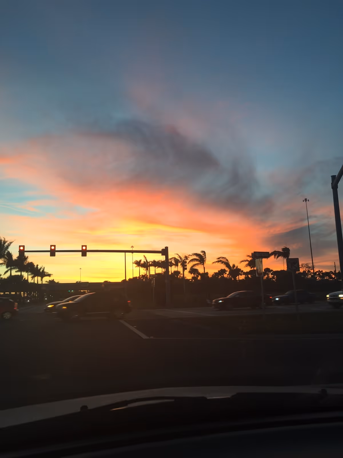 View of a sunset with vibrant orange, yellow, and blue colors in the sky above a street intersection with traffic lights and several cars. Palm trees line the horizon under the colorful sky.