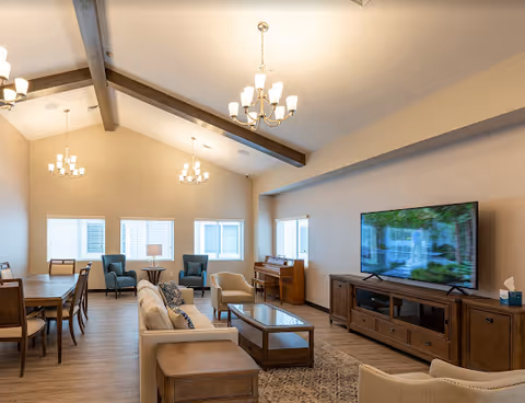 A spacious and well-lit living room with vaulted ceilings and exposed wooden beams. The room features a large flat-screen TV on a wooden media console, a beige sofa with patterned cushions, a glass coffee table, two armchairs near windows with blinds, a wooden piano, and a dining table with chairs. Multiple chandeliers hang from the ceiling, and the floor is covered with wood-style flooring and a patterned rug.