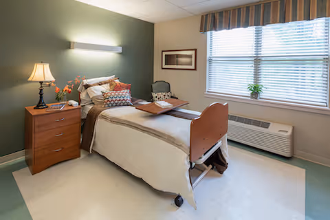 A neatly arranged bedroom in a healthcare facility featuring a single hospital-style bed with a wooden headboard and footboard, a bedside table with a lamp and flowers, a chair, a framed picture on the wall, and a large window with blinds and a striped valance. The room has a green accent wall and light-colored flooring.