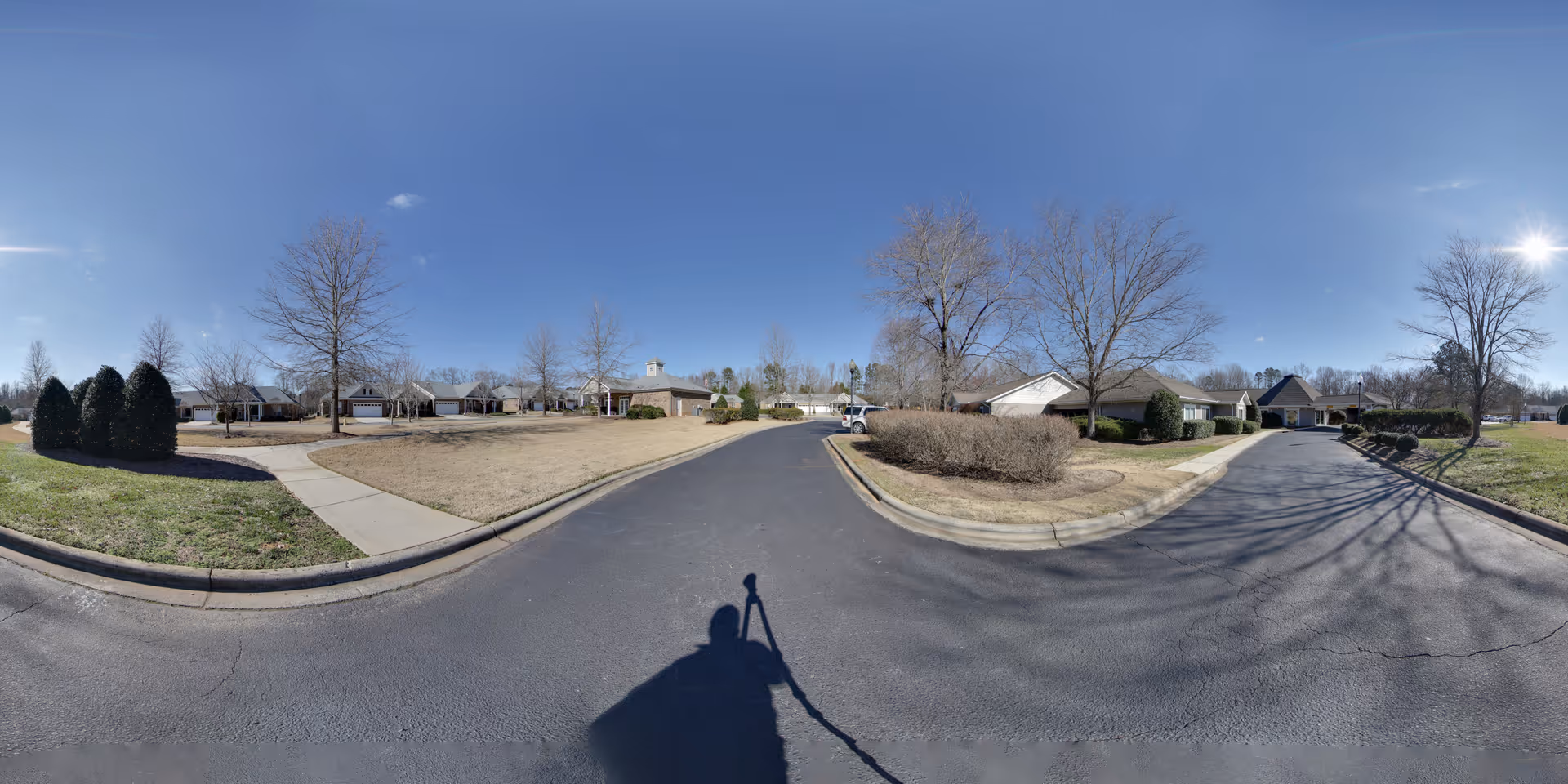 Wide panoramic view of a senior living community street with single-story houses, leafless trees, trimmed bushes, sidewalks, and a clear blue sky with the sun shining.