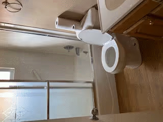 A clean bathroom featuring a white toilet, a bathtub with a sliding glass door, a wall-mounted showerhead, and a wooden vanity with a countertop. The floor has a wood-like finish, and the walls are painted light beige.