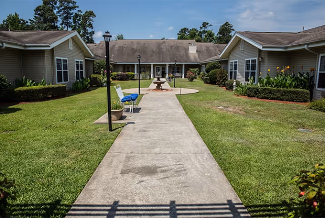 A concrete walkway leads through a well-maintained grassy courtyard with shrubs and flowers, flanked by single-story beige buildings with white trim. There are two blue chairs and a lamp post along the path, and a decorative fountain is visible in the center of the courtyard under a partly cloudy sky.