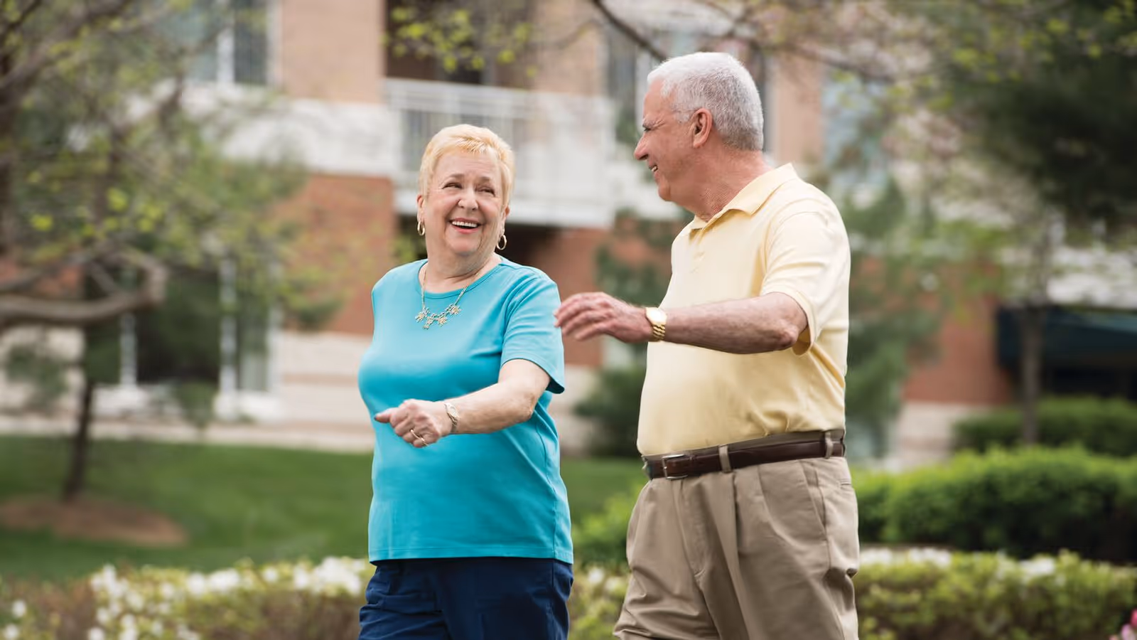An elderly man and woman walking outdoors in a garden area, smiling and engaging in conversation with a building in the background.