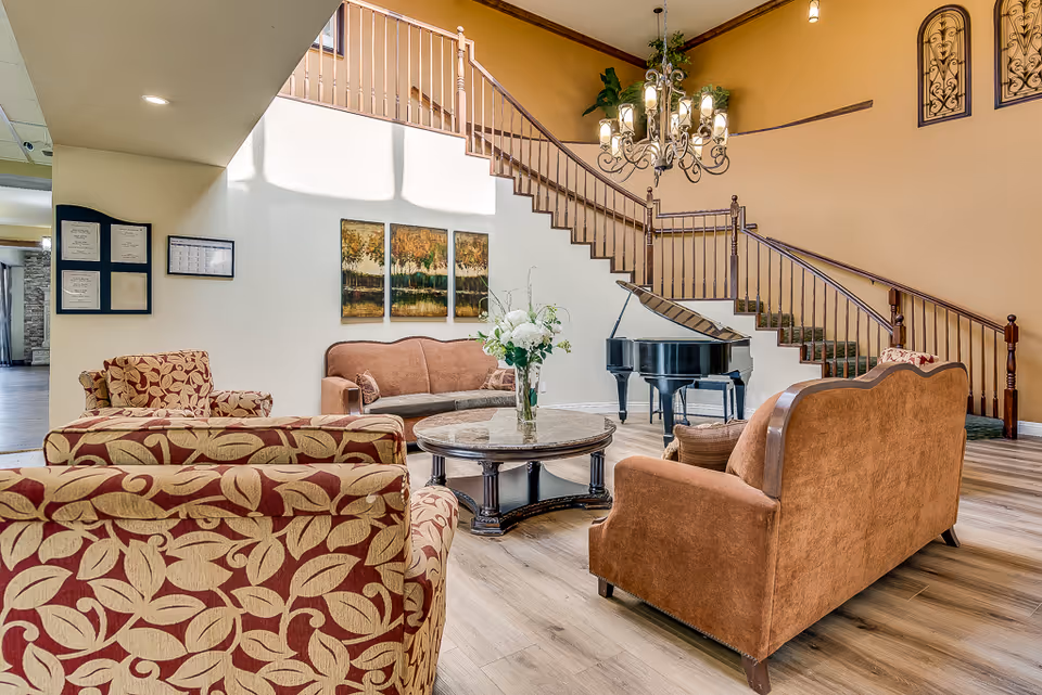 A spacious senior living facility common area with comfortable upholstered chairs and sofas arranged around a round wooden coffee table with a vase of white flowers. A grand piano is positioned near a staircase with wooden railings. The walls are decorated with framed artwork and ornamental wall hangings, and a chandelier hangs from the ceiling.
